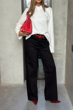 Person wearing a white shirt, black pants, and red shoes holding a red handbag against a gray wall.