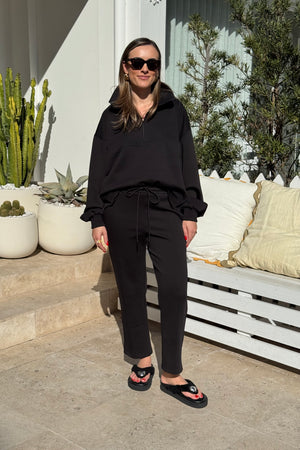 The image showcases a woman in a black half-zip sweatshirt and drawstring pants, complemented by black platform sandals and sunglasses. She is standing outdoors on a patio or garden area next to potted plants and a white wooden bench. The dominant colors are black and white, creating a simple and relaxed atmosphere. The composition is a centered, full shot highlighting the woman and her attire.