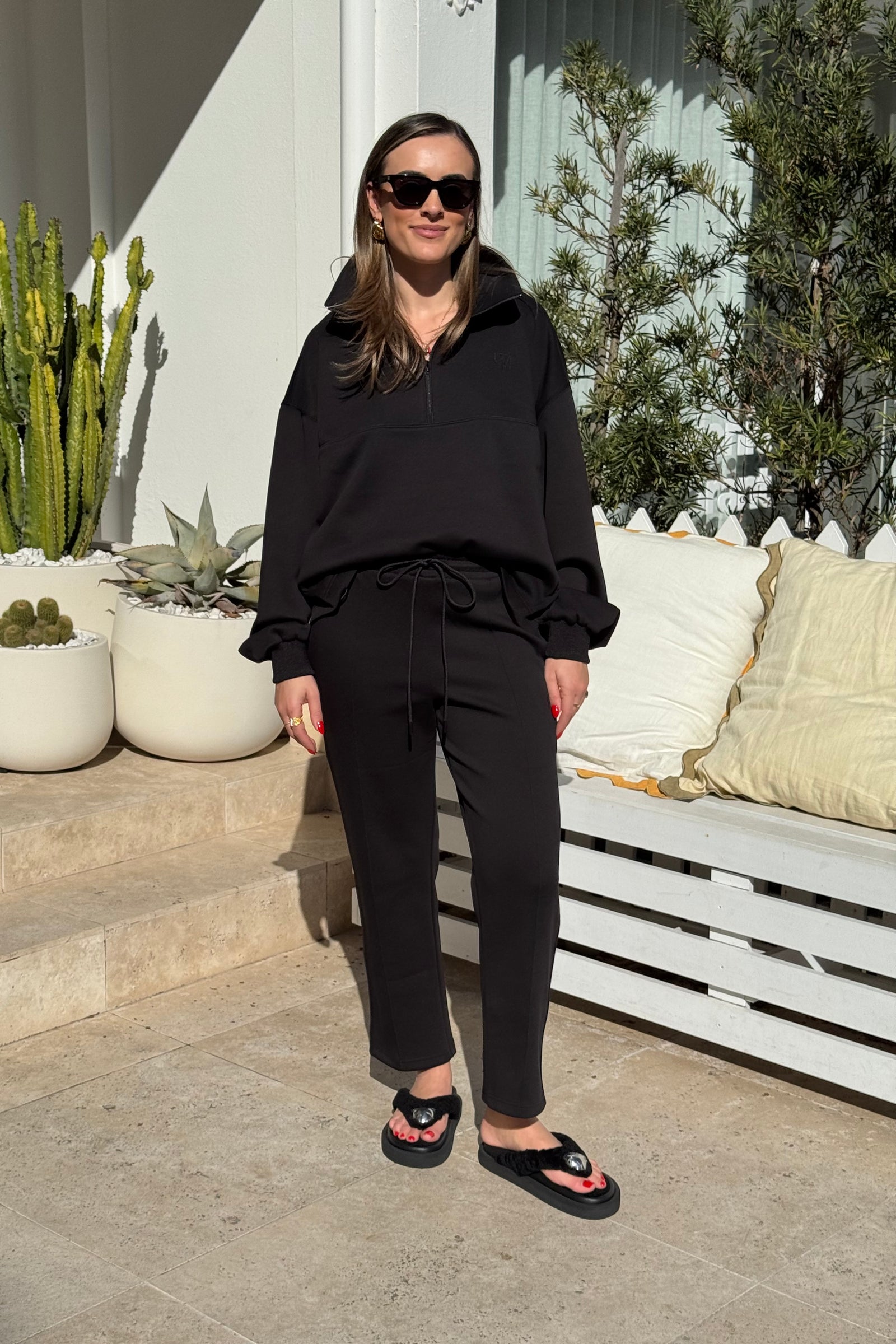 The image features a woman modeling a black athleisure outfit consisting of a half-zip sweatshirt and drawstring pants with a small logo, paired with black platform flip-flops. She is standing on a tiled patio with a white wooden bench and building in the background, presenting a casual yet stylish look.