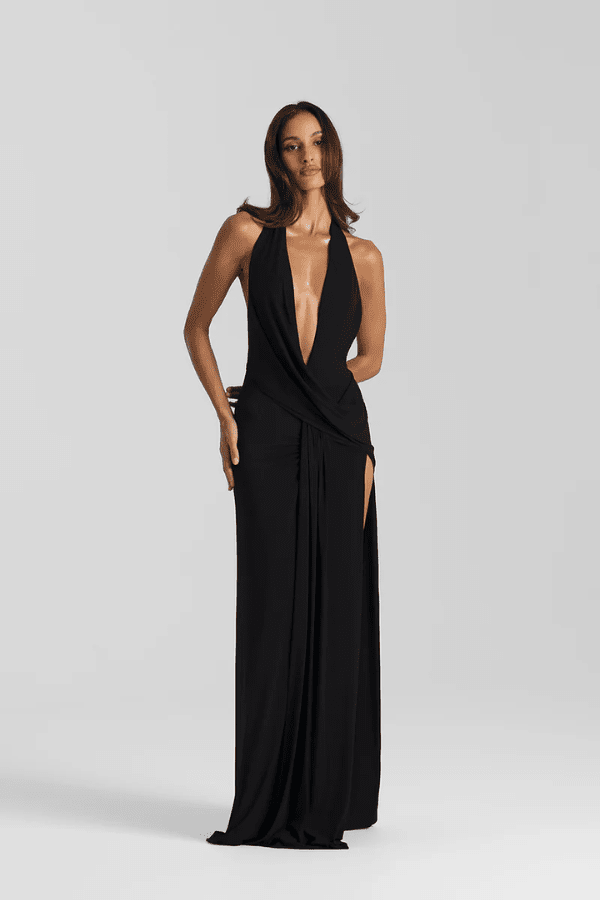 Winona Gown Black by Natalie Rolt, halter neckline, asymmetric draped bust overlay, open back, side split, gathered waterfall skirt, full-length hem, elevated jersey. Dry clean only.