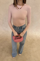 The Polly Long Sleeve top in pink. Woman in a pink top and white pants standing outside a store with plants and a bench in the background.