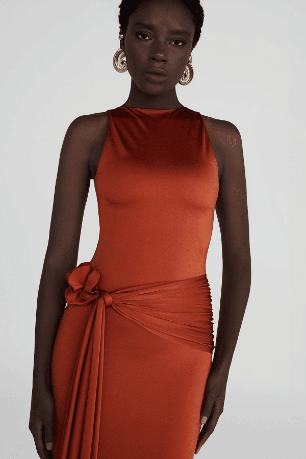 Tirso Terracotta by Maygel Coronel – Long dress with round neck, open back, ruched sash, and 3D flowers for elegant look.