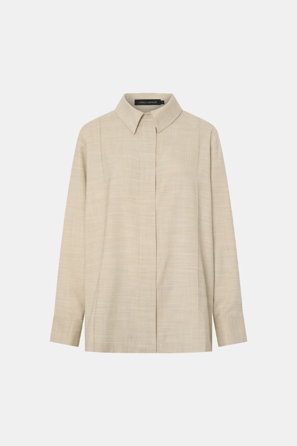 The image shows a woman modeling a pale beige long-sleeve button-down shirt and drawstring pants. She is wearing gold earrings, a gold ring, and a gold bracelet. The photo seems to be taken in a shop or studio, with a cactus in a white pot visible in the background. The composition is a medium shot focusing on the outfit.