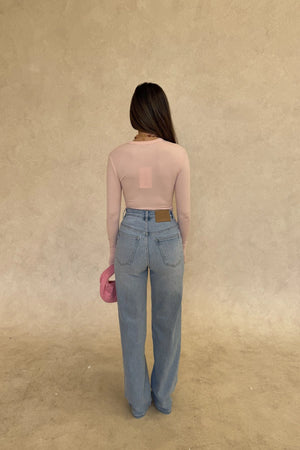 The Polly Long Sleeve top in pink. Woman in a pink top and white pants standing outside a store with plants and a bench in the background.