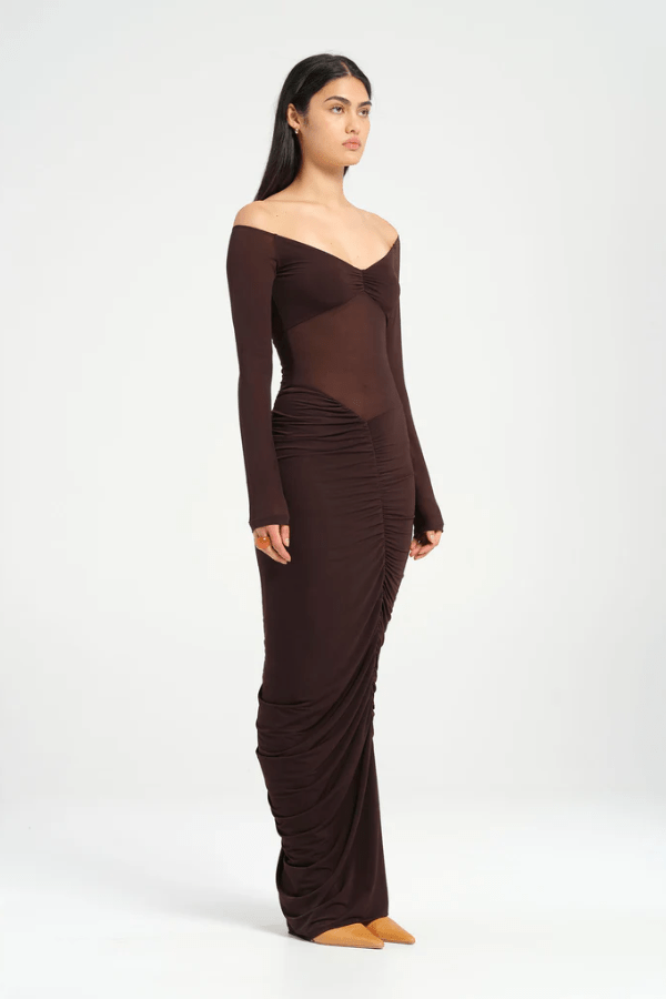 Wilma Maxi Dress Chocolate by Benni, featuring an off-shoulder V-neckline, full-length sleeves, sheer waist, and asymmetrical drape for a sculpted silhouette. Ideal for statement evenings.