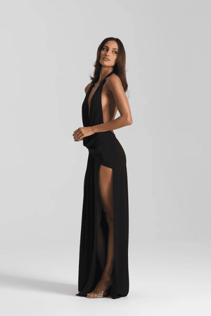 Winona Gown Black by Natalie Rolt, halter neckline, asymmetric draped bust overlay, open back, side split, gathered waterfall skirt, full-length hem, elevated jersey. Dry clean only.