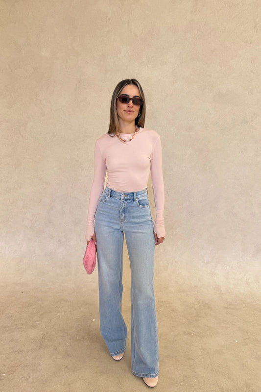 The Polly Long Sleeve top in pink. Woman in a pink top and white pants standing outside a store with plants and a bench in the background.