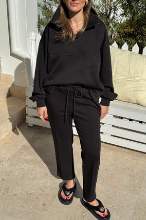 The image features a woman modeling a black athleisure outfit consisting of a half-zip sweatshirt and drawstring pants with a small logo, paired with black platform flip-flops. She is standing on a tiled patio with a white wooden bench and building in the background, presenting a casual yet stylish look.