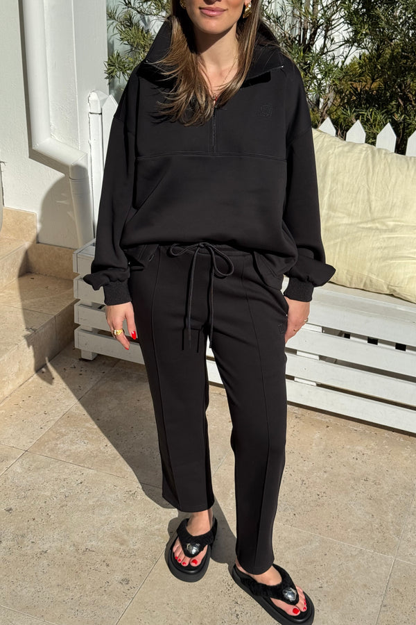 The image features a woman modeling a black athleisure outfit consisting of a half-zip sweatshirt and drawstring pants with a small logo, paired with black platform flip-flops. She is standing on a tiled patio with a white wooden bench and building in the background, presenting a casual yet stylish look.