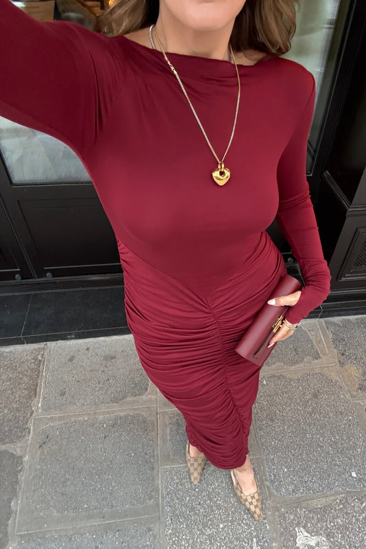 The image is a high-angle shot of a woman showcasing her outfit, which includes a burgundy, long-sleeved dress, a gold heart pendant necklace, a burgundy clutch, and Gucci high heels. The setting appears to be outdoors in front of a dark door. The colors are predominantly burgundy, gold, white, light brown, and gray.