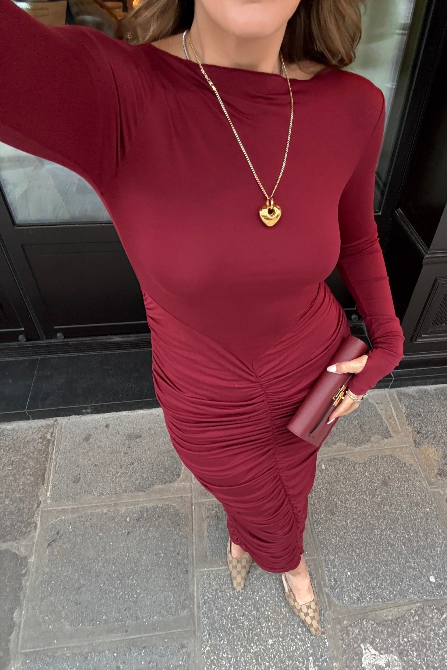 The image is a high-angle shot of a woman showcasing her outfit, which includes a burgundy, long-sleeved dress, a gold heart pendant necklace, a burgundy clutch, and Gucci high heels. The setting appears to be outdoors in front of a dark door. The colors are predominantly burgundy, gold, white, light brown, and gray.