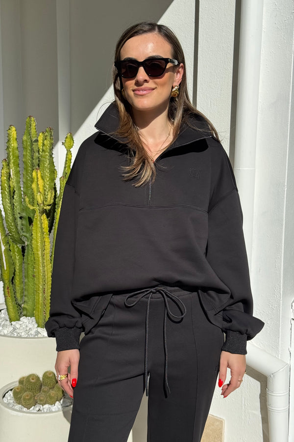 The image shows a woman outdoors, standing next to a white wall with a cactus in a white pot. She's wearing a black oversized sweatshirt with a logo, black pants with a drawstring, sunglasses, gold earrings, a gold necklace, and gold rings. The primary color scheme is black, gold, red, and green against a white backdrop, creating a modern and stylish aesthetic. She appears relaxed and confident.