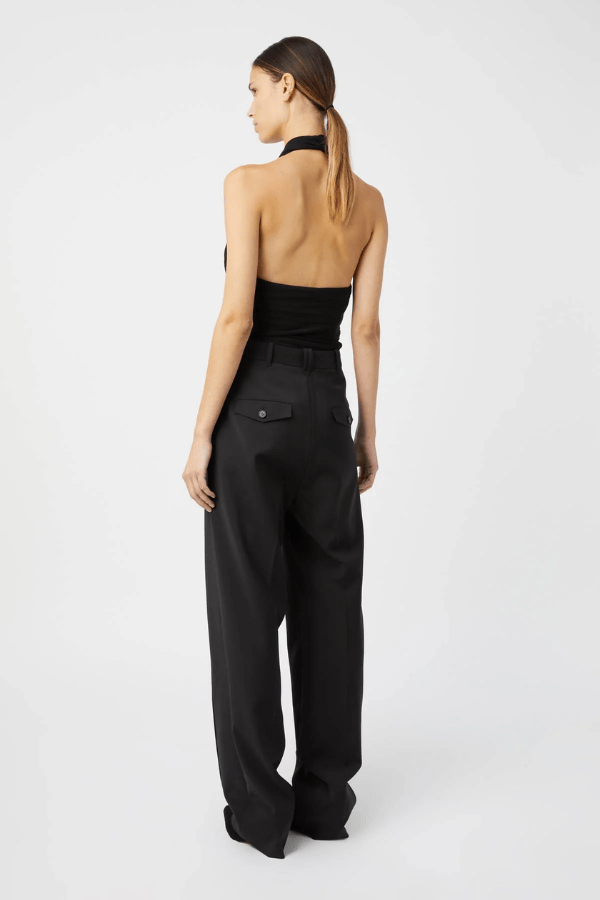 The image features a smiling woman wearing a black halter top and dress pants. She is also wearing gold hoop earrings and gold bracelets. The halter top has a unique square connector with illegible text. The photo is a medium shot, taken indoors against a plain white wall. The overall impression is one of simple elegance.