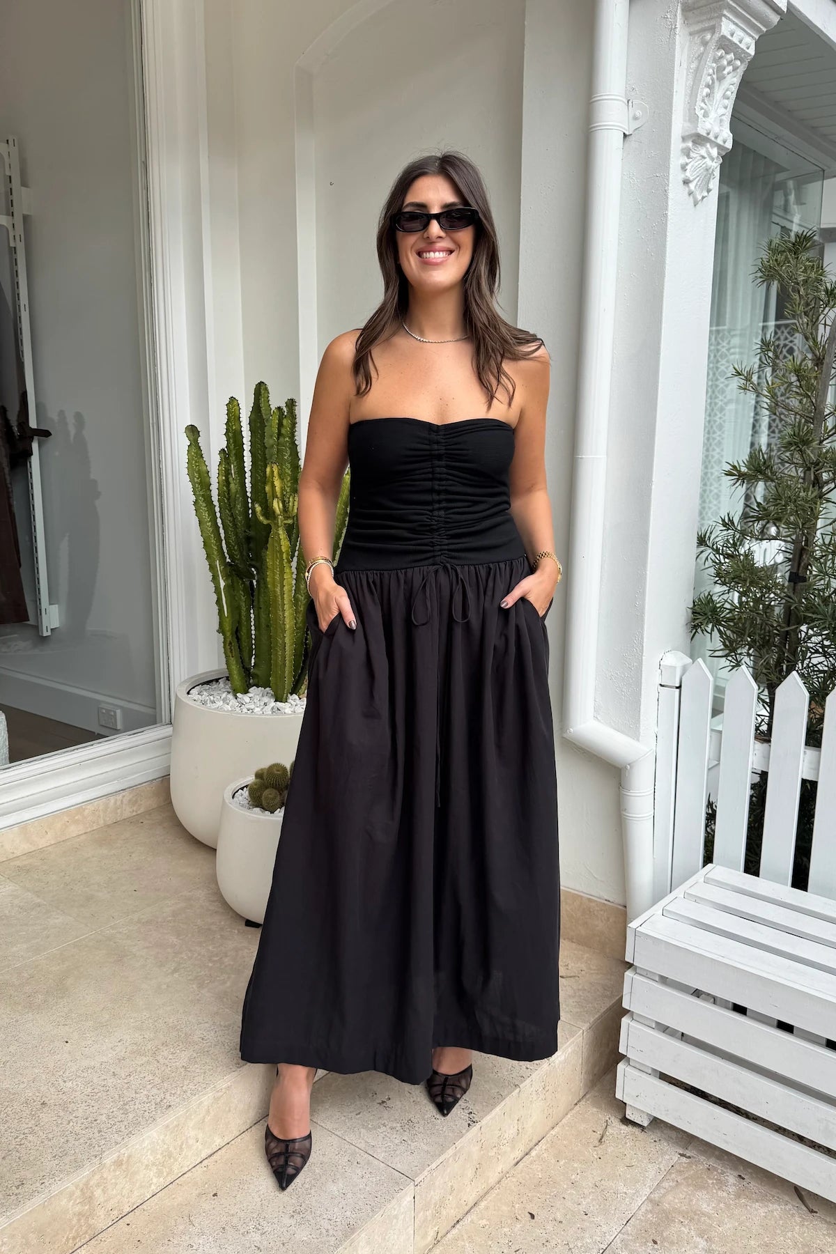 The image shows a smiling woman with long brown hair, wearing a black strapless top, a long black skirt, and black pointed-toe heels. She also has sunglasses, a necklace, and bracelets on. She is standing on what appears to be a storefront patio near a white wall beside a white bench and potted cacti. The overall scene is bright and well-lit, giving off a relaxed and confident vibe.