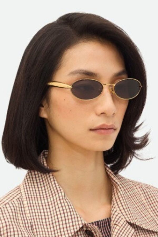 Bottega Veneta BV1386S sunglasses, oval gold frame with sardine-patterned temples and grey lenses. A playful yet chic statement piece.
