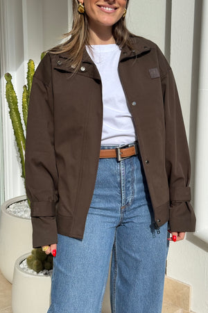The image features a smiling woman wearing a brown jacket, a white t-shirt, and blue jeans, accessorized with a brown leather belt, gold earrings, and a gold ring. Her fingernails are painted bright red. The photo is taken in a brightly lit area with a white wall and a potted cactus in the background. The woman's attire suggests a casual yet stylish look. There is a small logo on the jacket that is unidentifiable.