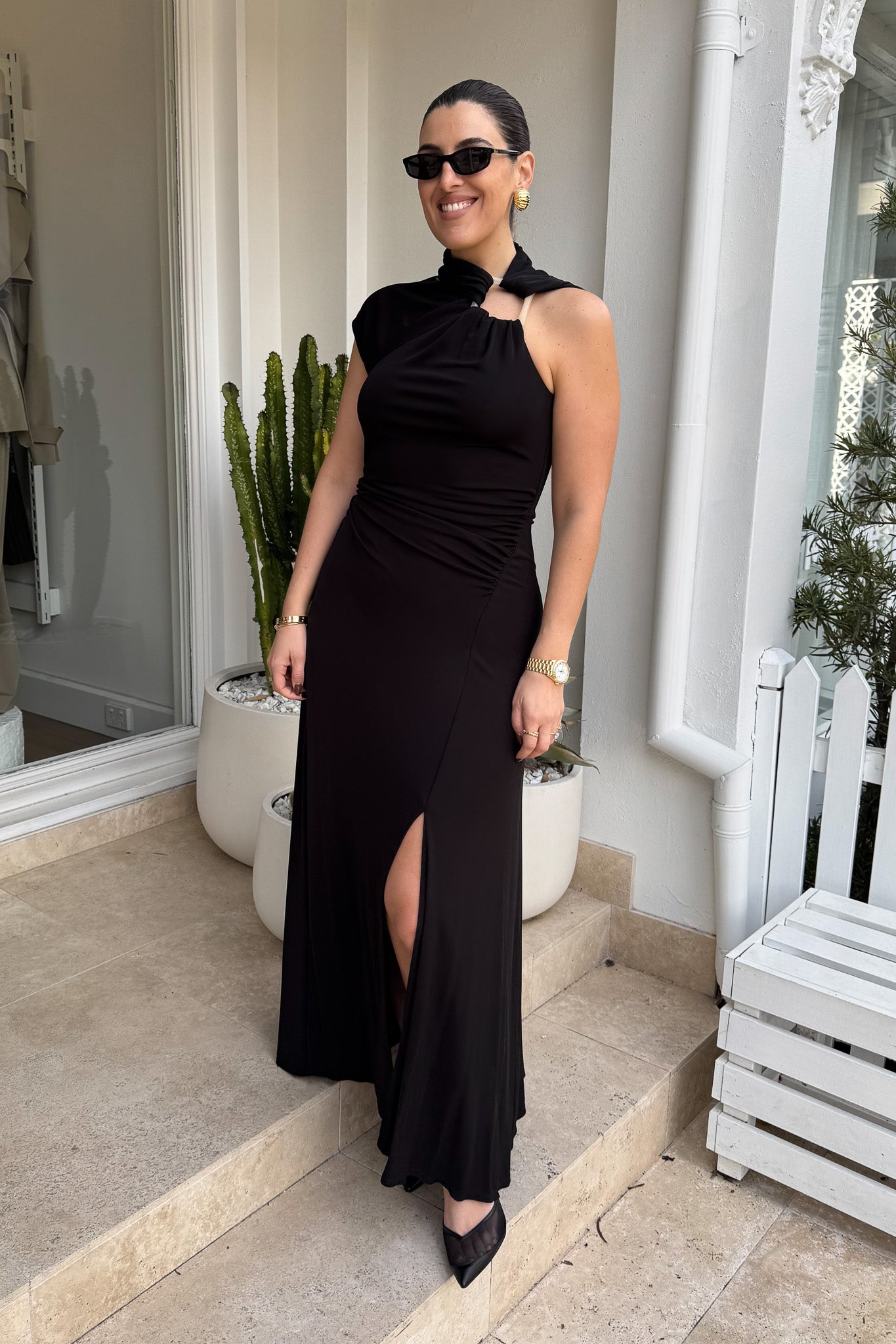 The image shows a smiling woman in a black maxi dress with a side slit and black heels, standing on stone steps outside a building with a white exterior. She is accessorized with black sunglasses, gold hoop earrings, a gold watch, and a gold bracelet. To either side of her are a white planter with a cactus and a white planter with a green leafy plant, and to the right is a white bench.