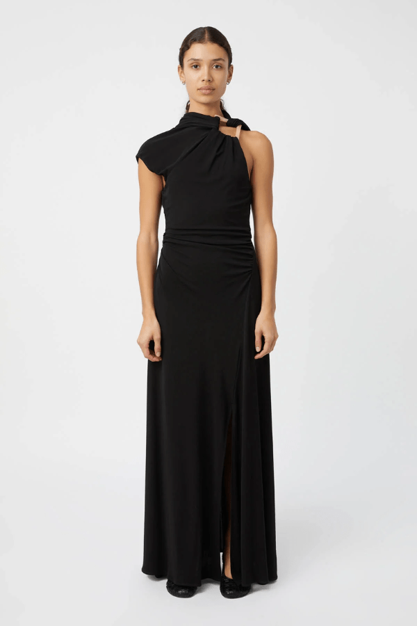 The image shows a smiling woman in a black maxi dress with a side slit and black heels, standing on stone steps outside a building with a white exterior. She is accessorized with black sunglasses, gold hoop earrings, a gold watch, and a gold bracelet. To either side of her are a white planter with a cactus and a white planter with a green leafy plant, and to the right is a white bench.