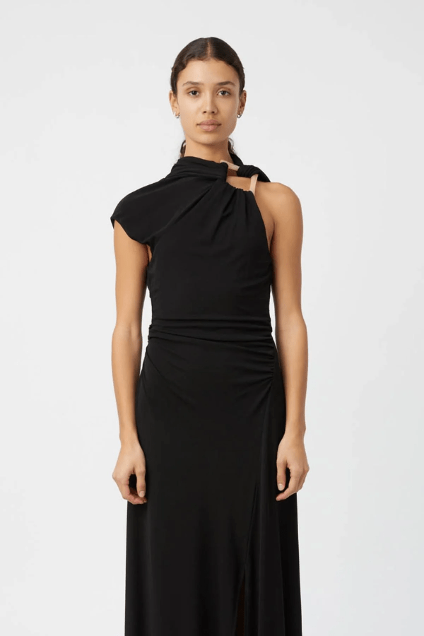 The image shows a smiling woman in a black maxi dress with a side slit and black heels, standing on stone steps outside a building with a white exterior. She is accessorized with black sunglasses, gold hoop earrings, a gold watch, and a gold bracelet. To either side of her are a white planter with a cactus and a white planter with a green leafy plant, and to the right is a white bench.