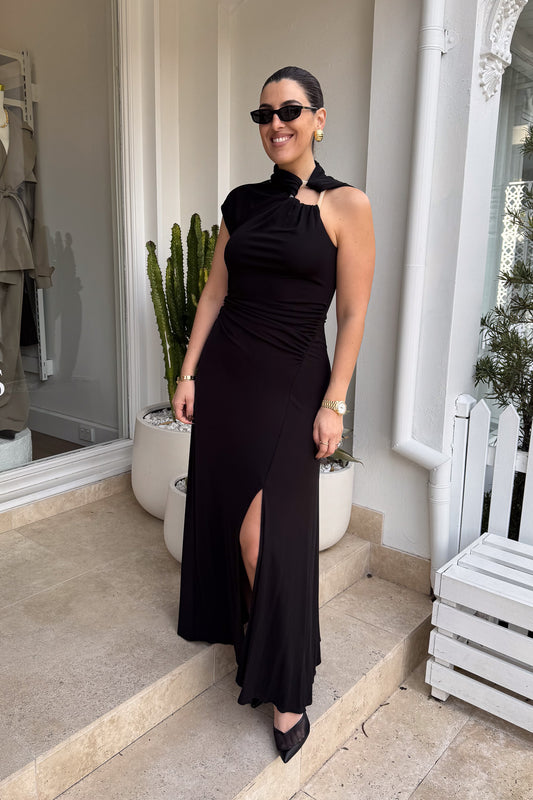 The image shows a smiling woman in a black maxi dress with a side slit and black heels, standing on stone steps outside a building with a white exterior. She is accessorized with black sunglasses, gold hoop earrings, a gold watch, and a gold bracelet. To either side of her are a white planter with a cactus and a white planter with a green leafy plant, and to the right is a white bench.