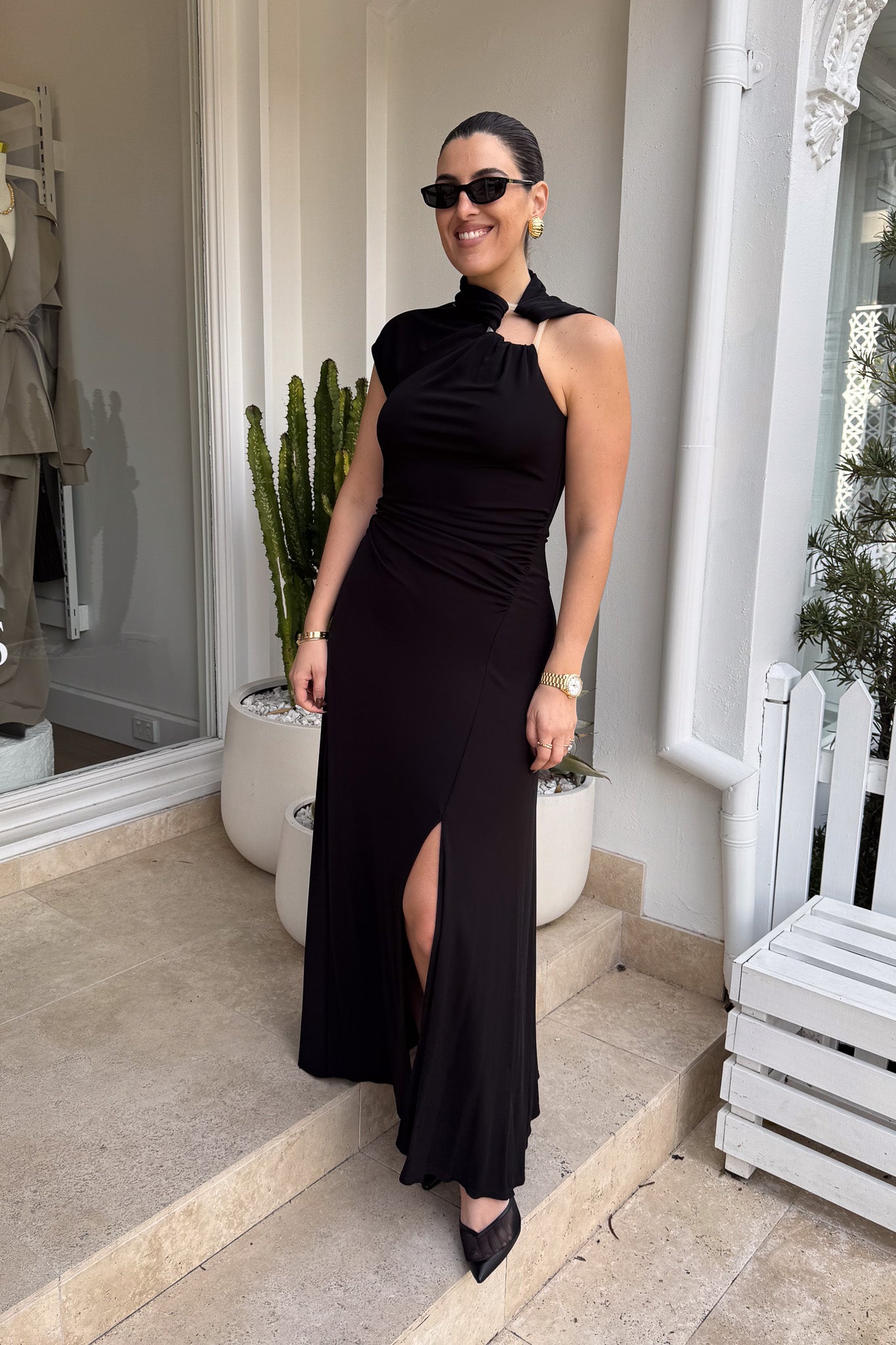 The image shows a smiling woman in a black maxi dress with a side slit and black heels, standing on stone steps outside a building with a white exterior. She is accessorized with black sunglasses, gold hoop earrings, a gold watch, and a gold bracelet. To either side of her are a white planter with a cactus and a white planter with a green leafy plant, and to the right is a white bench.