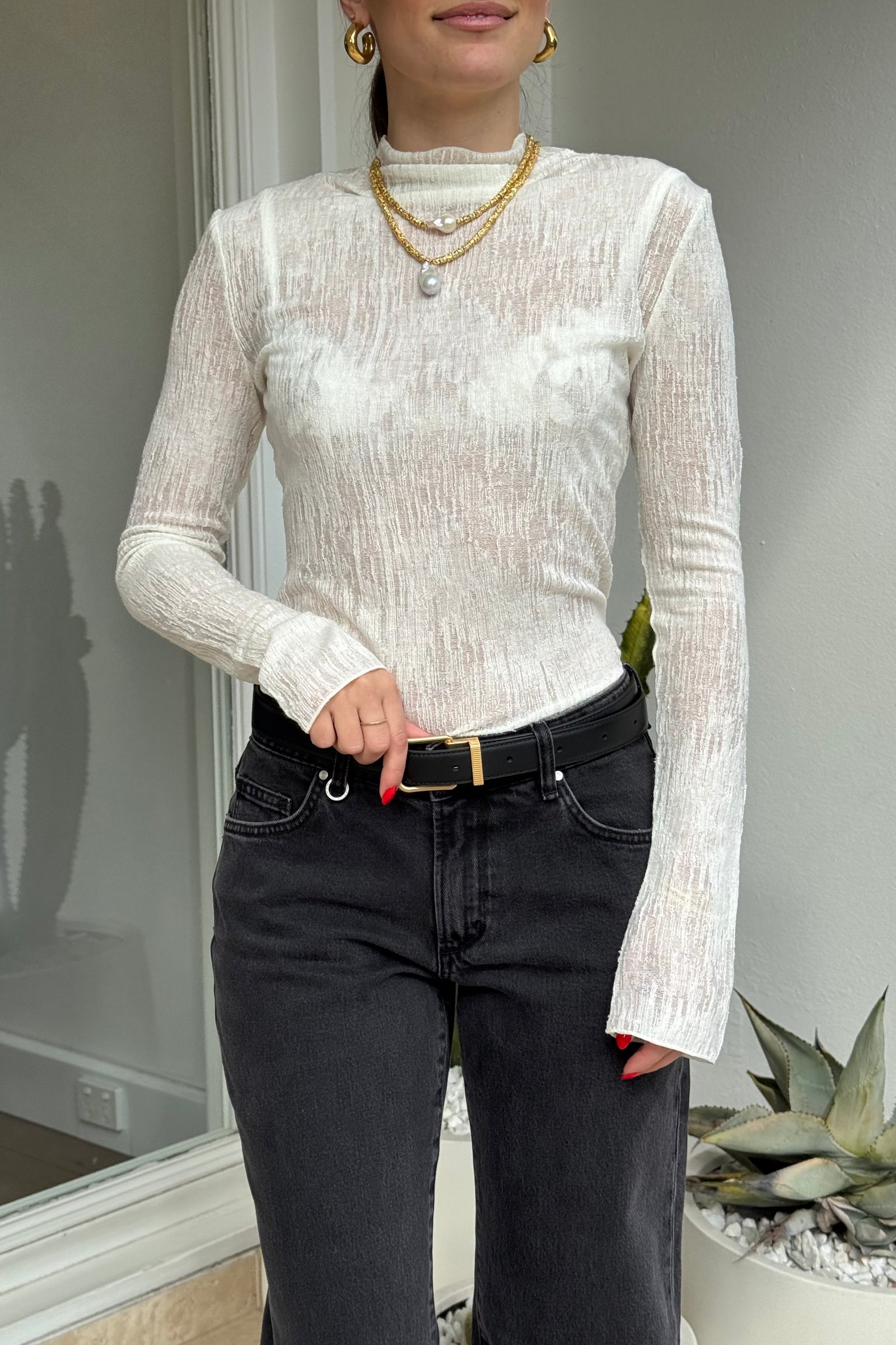 The image features a woman modeling a white textured long-sleeved top paired with dark denim jeans and a black leather belt with a gold buckle. She is accessorized with gold hoop earrings and a two-strand gold necklace featuring pearl pendants. The shot is indoors, with natural lighting, and includes a potted succulent plant in the background.