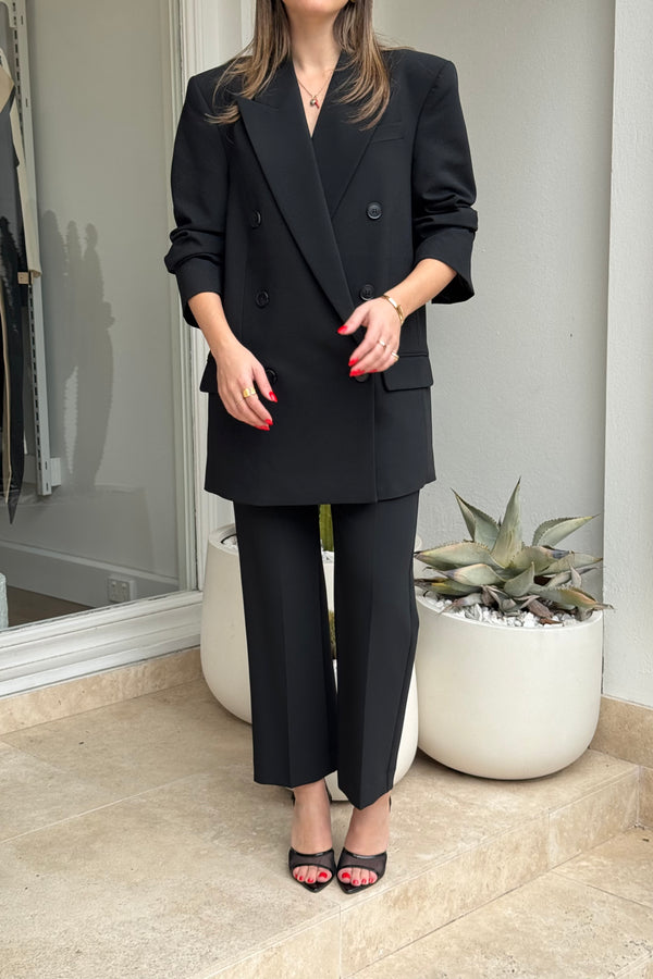 The image shows a woman standing in front of a building entrance or storefront, wearing a black double-breasted blazer, black pants, and black heeled sandals. She has medium-length brown hair and red nail polish, accessorized with gold jewelry. The setting includes two large, white planters, one containing an agave plant. The color scheme is primarily black and white, with neutral earth tones from the travertine tile floor.