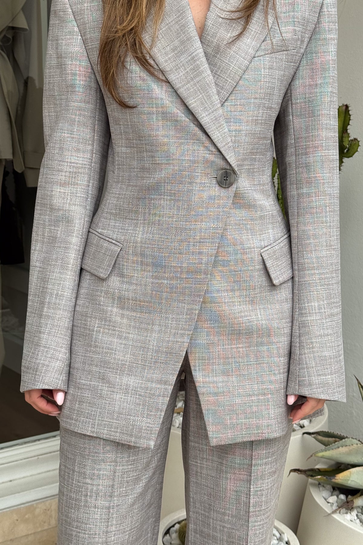 The image showcases a person wearing a gray plaid blazer with a one-button closure and wide-leg pants. The setting includes a cactus in a white pot, adding a modern touch to the scene. The shot focuses on the details and fit of the clothing, with the gray tones accented by subtle colors in the plaid pattern.