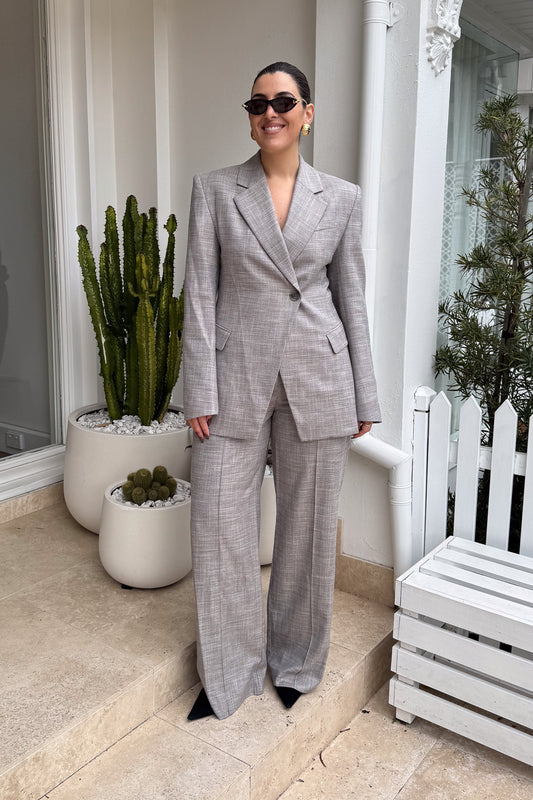 A woman stands confidently on a tiled patio in front of a white building, wearing a stylish gray suit with wide-leg pants and black pointed-toe heels. She accessorizes with sunglasses and gold earrings, smiling at the camera. Potted cacti flank her, and a white wooden bench sits to her right. The photo is a well-lit, full shot focusing on the subject's fashion and poised demeanor.