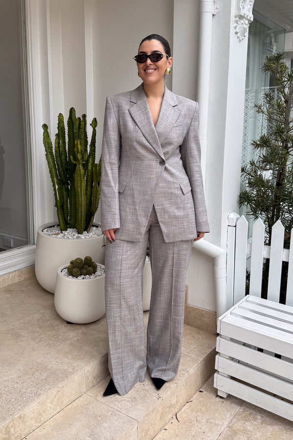 A woman stands confidently on a tiled patio in front of a white building, wearing a stylish gray suit with wide-leg pants and black pointed-toe heels. She accessorizes with sunglasses and gold earrings, smiling at the camera. Potted cacti flank her, and a white wooden bench sits to her right. The photo is a well-lit, full shot focusing on the subject's fashion and poised demeanor.