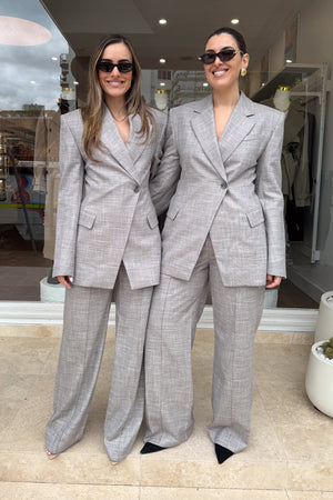 Two women stand side-by-side in front of a storefront, both wearing matching grey suits, sunglasses, and smiles. The suits feature an asymmetrical button closure and wide-leg pants. The woman on the left has long brown hair, while the woman on the right has dark hair pulled back and is wearing large gold hoop earrings. The scene is well-lit and the subjects are centered.