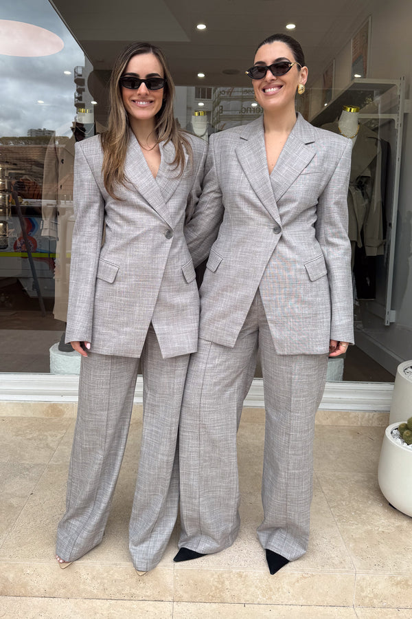 Two women stand side-by-side in front of a storefront, both wearing matching grey suits, sunglasses, and smiles. The suits feature an asymmetrical button closure and wide-leg pants. The woman on the left has long brown hair, while the woman on the right has dark hair pulled back and is wearing large gold hoop earrings. The scene is well-lit and the subjects are centered.