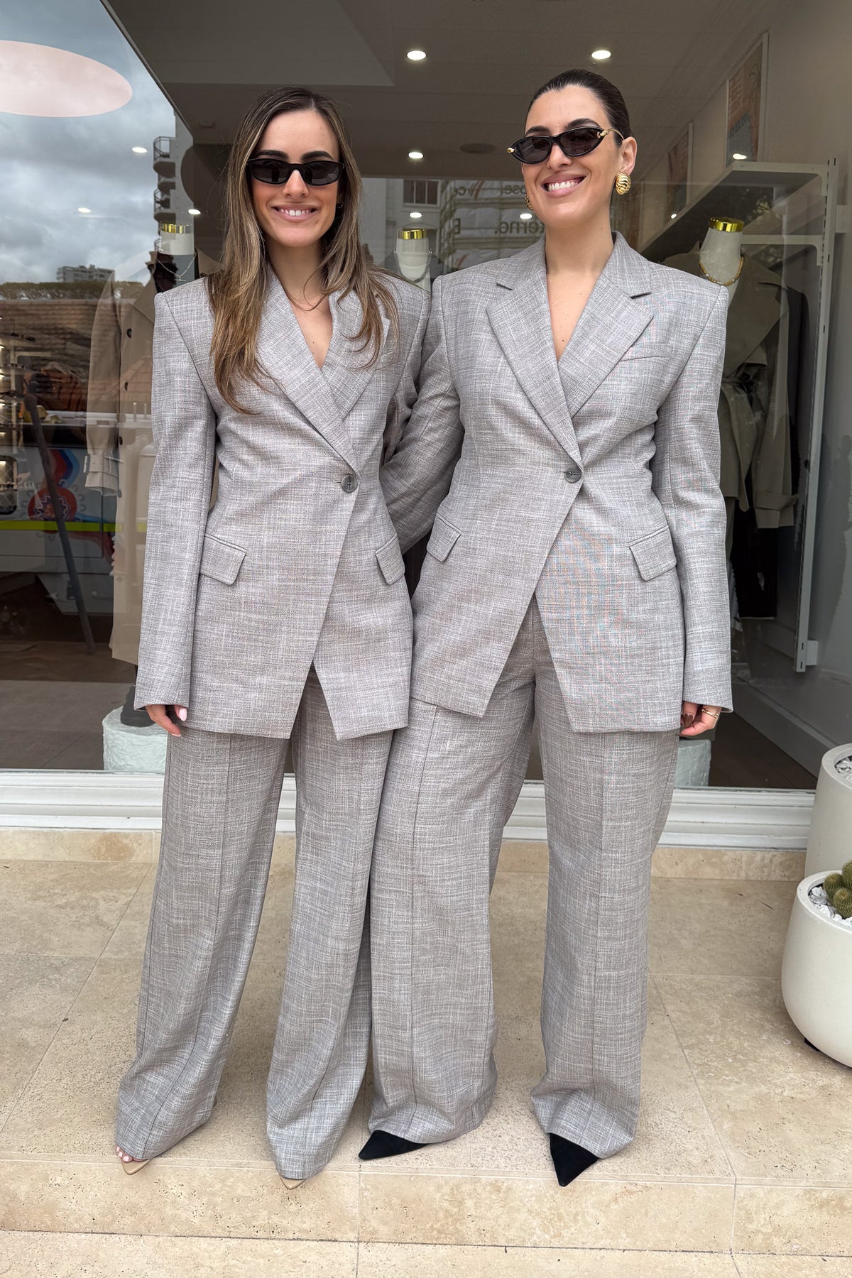 Two women stand side-by-side in front of a storefront, both wearing matching grey suits, sunglasses, and smiles. The suits feature an asymmetrical button closure and wide-leg pants. The woman on the left has long brown hair, while the woman on the right has dark hair pulled back and is wearing large gold hoop earrings. The scene is well-lit and the subjects are centered.