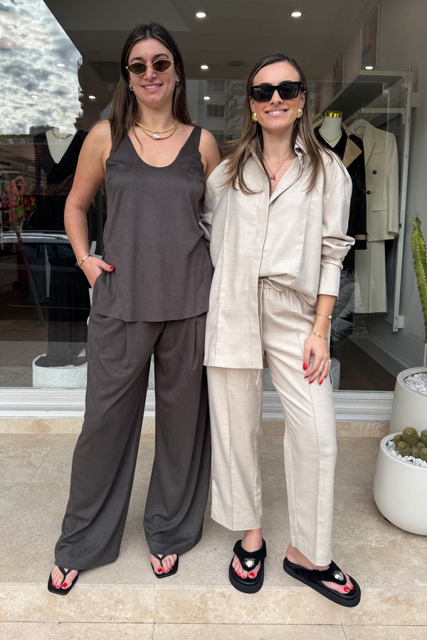 Two women are posing in front of a building with a storefront. The woman on the left is wearing a gray tank top with loose-fitting gray pants, while the woman on the right is wearing a beige long-sleeved shirt and drawstring pants. Both are wearing sunglasses, necklaces, bracelets, and flip-flops, with red nail polish on their toes and fingers. The storefront window displays clothing on mannequins. Cacti in planters are positioned near the entrance.