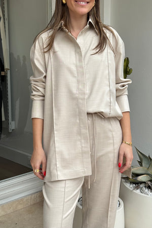 The image shows a woman modeling a pale beige long-sleeve button-down shirt and drawstring pants. She is wearing gold earrings, a gold ring, and a gold bracelet. The photo seems to be taken in a shop or studio, with a cactus in a white pot visible in the background. The composition is a medium shot focusing on the outfit.