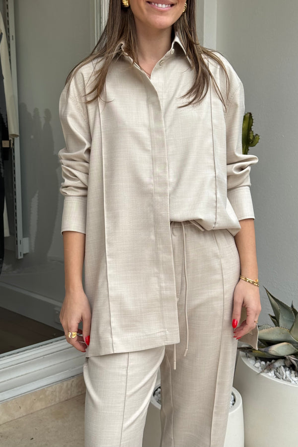 The image shows a woman modeling a pale beige long-sleeve button-down shirt and drawstring pants. She is wearing gold earrings, a gold ring, and a gold bracelet. The photo seems to be taken in a shop or studio, with a cactus in a white pot visible in the background. The composition is a medium shot focusing on the outfit.