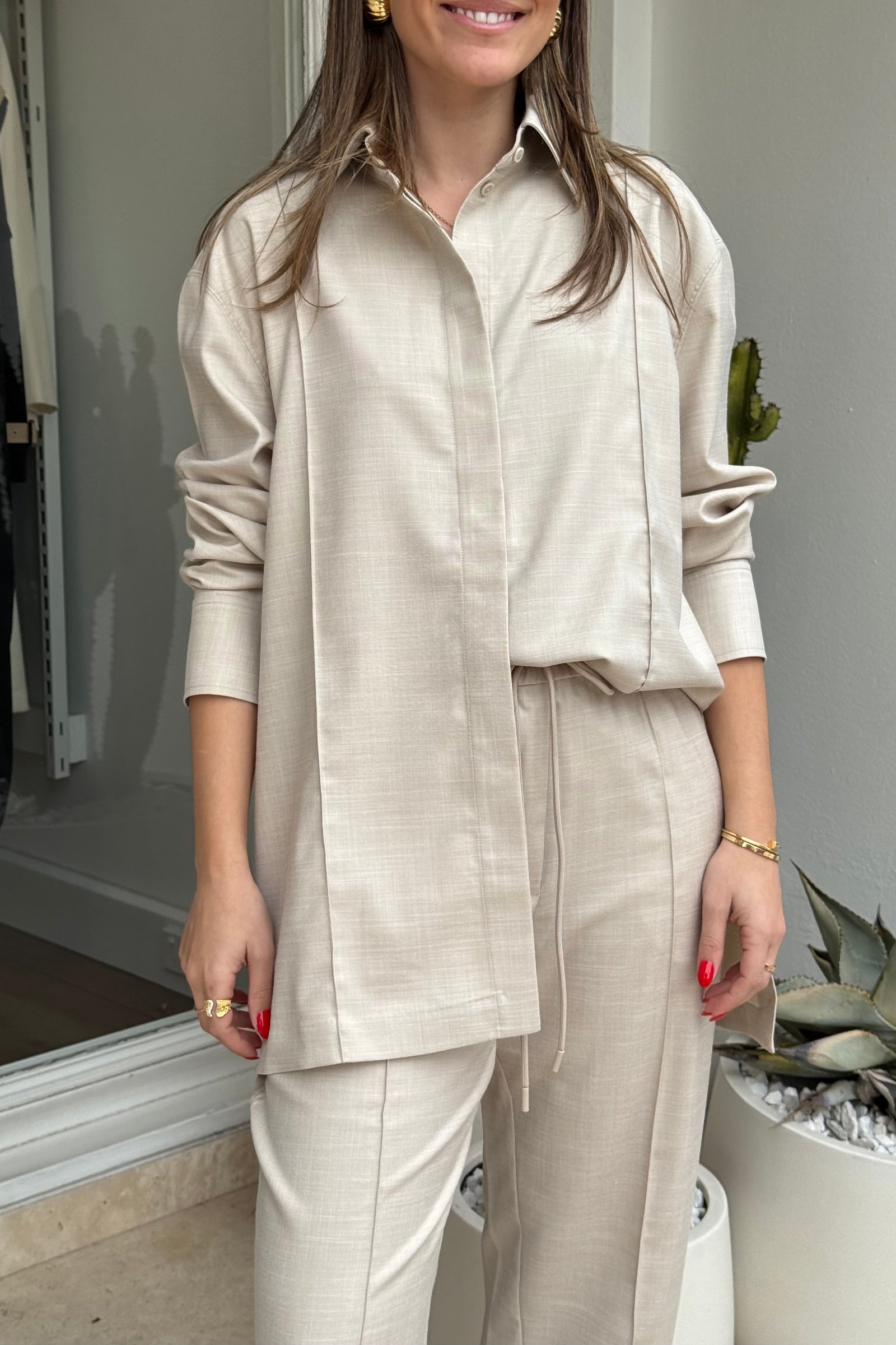 The image features a woman modeling a light beige two-piece shirt and pants outfit outside of a store, as can be seen with the "MS" text on the store window, accessorized with black platform sandals, sunglasses, and gold jewelry. The background includes potted plants, providing a natural element to the fashion-oriented scene. The woman stands confidently, showcasing the clothing in an outdoor setting.