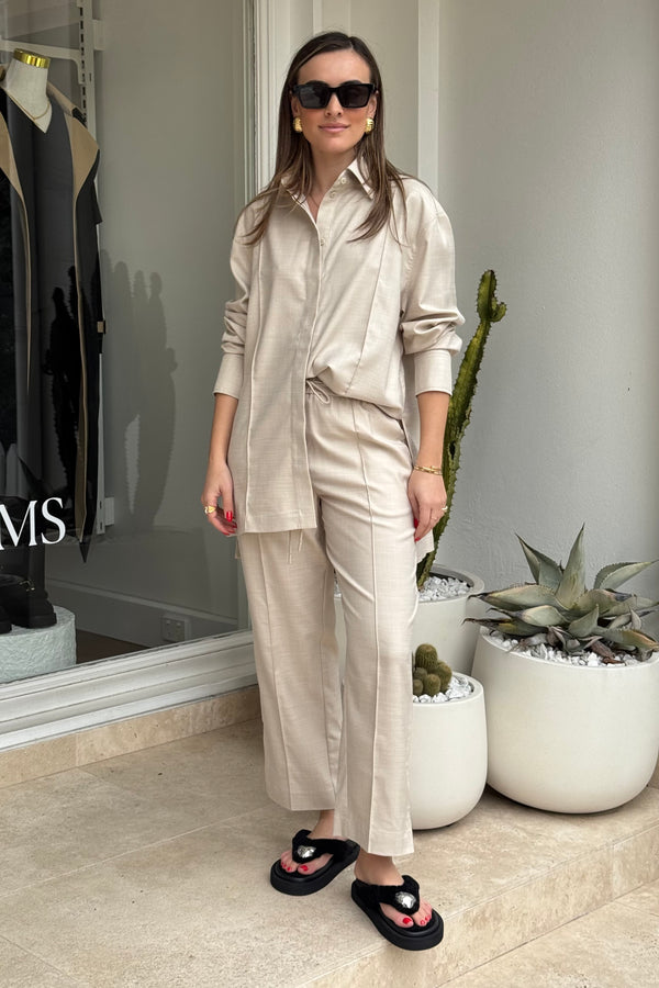 The image features a woman modeling a light beige two-piece shirt and pants outfit outside of a store, as can be seen with the "MS" text on the store window, accessorized with black platform sandals, sunglasses, and gold jewelry. The background includes potted plants, providing a natural element to the fashion-oriented scene. The woman stands confidently, showcasing the clothing in an outdoor setting.