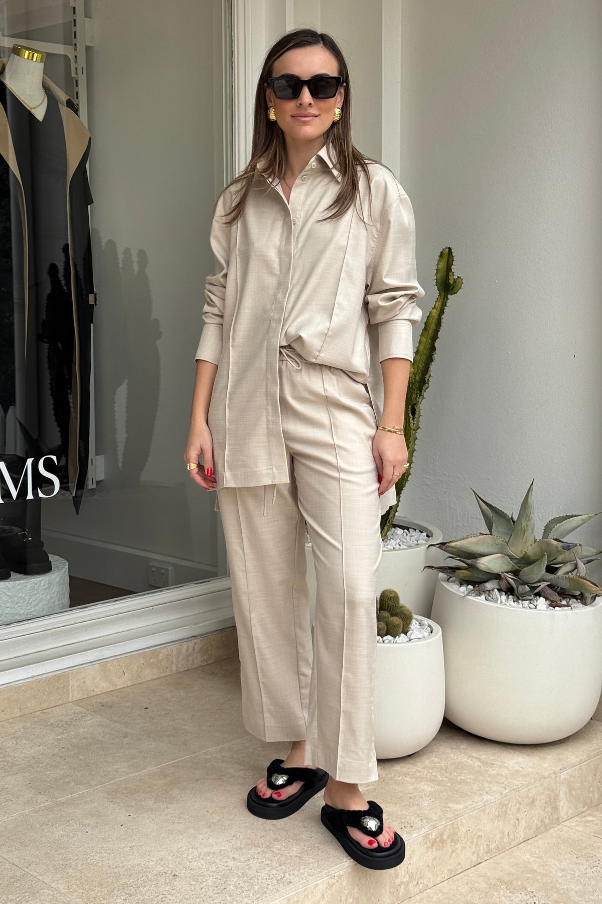 The image features a woman modeling a light beige two-piece shirt and pants outfit outside of a store, as can be seen with the "MS" text on the store window, accessorized with black platform sandals, sunglasses, and gold jewelry. The background includes potted plants, providing a natural element to the fashion-oriented scene. The woman stands confidently, showcasing the clothing in an outdoor setting.