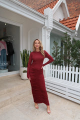 The image is a high-angle shot of a woman showcasing her outfit, which includes a burgundy, long-sleeved dress, a gold heart pendant necklace, a burgundy clutch, and Gucci high heels. The setting appears to be outdoors in front of a dark door. The colors are predominantly burgundy, gold, white, light brown, and gray.