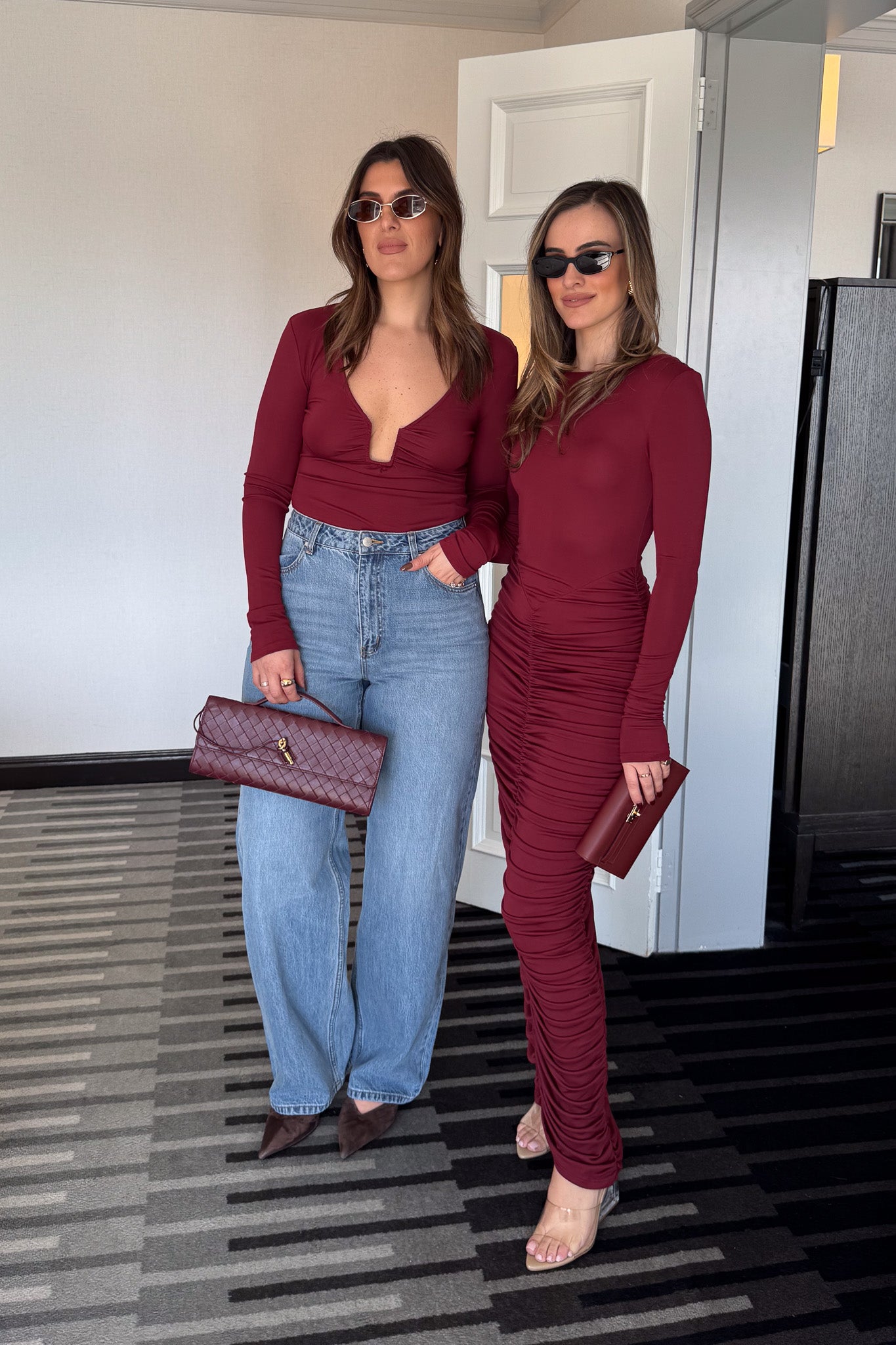 The image is a high-angle shot of a woman showcasing her outfit, which includes a burgundy, long-sleeved dress, a gold heart pendant necklace, a burgundy clutch, and Gucci high heels. The setting appears to be outdoors in front of a dark door. The colors are predominantly burgundy, gold, white, light brown, and gray.