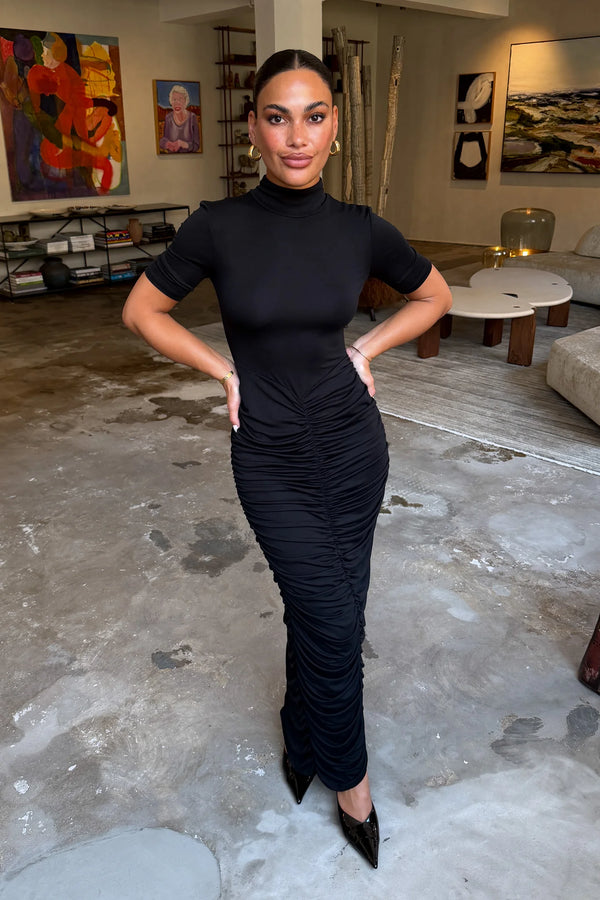 The Imana Draped Maxi Dress in Black by Sneaky Link features a flattering draped skirt, fitted upper, and soft modal stretch fabric for a feminine, timeless silhouette.