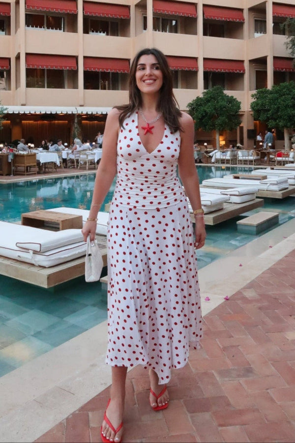 Woman in a white dress with red polka dots standing by a poolside with a hotel in the background.
