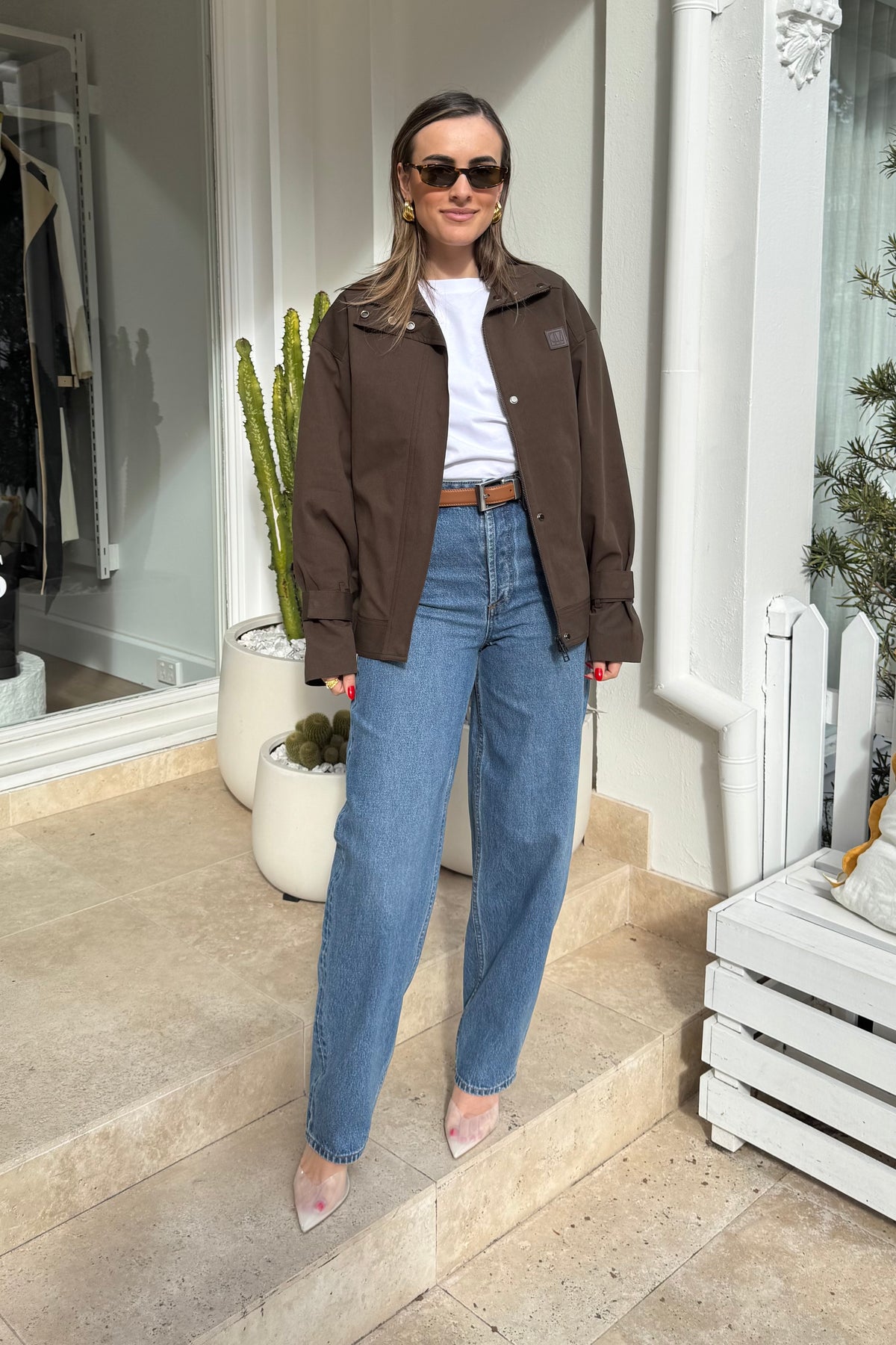 The image shows a woman standing on stone steps outdoors, possibly in front of a shop. She is wearing a brown jacket, white t-shirt, blue jeans with a brown belt, and clear heels, accessorized with gold hoop earrings and sunglasses. The setting includes potted cacti and white architectural details, contributing to a casual and well-lit scene.