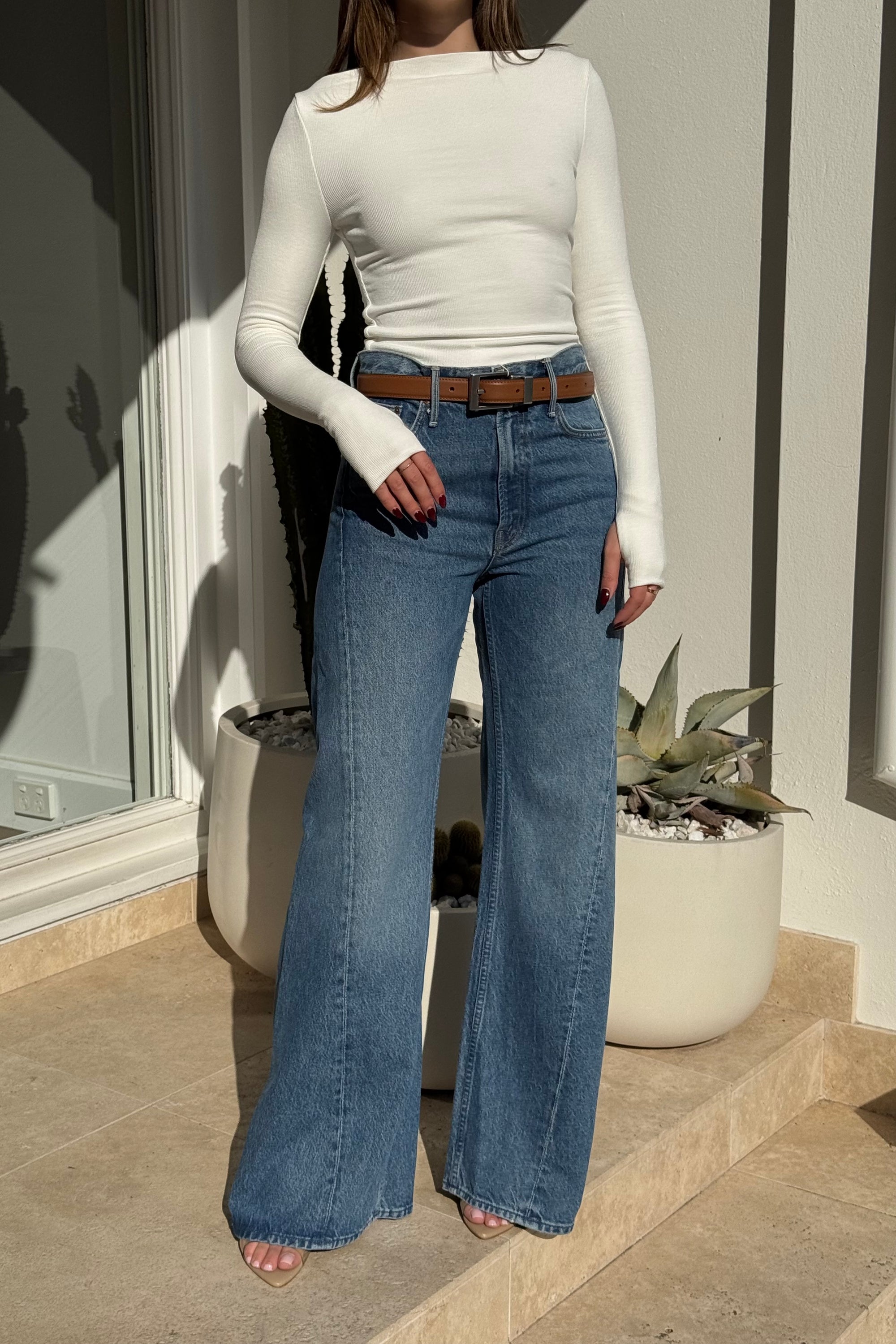 The image shows a person wearing a white long-sleeve shirt, blue wide-leg jeans with a brown leather belt, and beige sandals, standing outdoors on a tiled surface next to large white potted plants containing cacti and an agave. The setting is a brightly lit patio or entrance area.