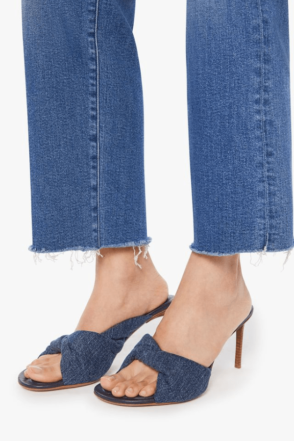 The image shows a smiling woman with dark hair wearing sunglasses, a long-sleeved brown top, blue jeans with a black belt, and flip-flops. She is standing outdoors or in an entrance area with a hand in her pocket, flanked by two large white planters containing plants. The scene is well-lit, and the color scheme is neutral with dominant colors of brown, blue, black, gold, and white.