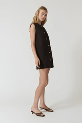 Nanda Dress Chocolate by Onte – Playful sleeveless mini shift with gold knot buttons, crew neck, welt pockets, and a flattering straight fit. Perfect for chic daytime events and casual outings.