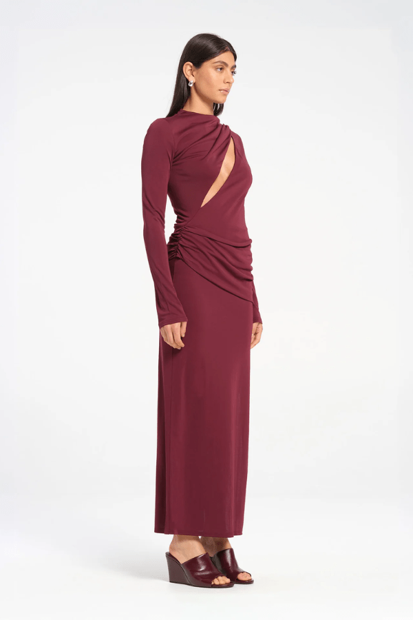 Sacha Mini Dress in Plum by Benni, crafted from stretch jersey with a high neckline, long sleeves, and angled chest cut-out. Draped waist sash and gathered skirt for dynamic texture.