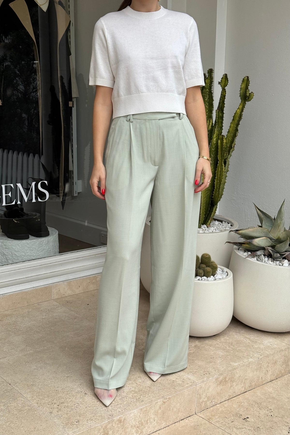 The image showcases a person modeling a white short-sleeved knit shirt and light green wide-leg pleated pants, accessorized with pointed-toe shoes and a gold bangle. The setting is in front of a store with the text "EMS" visible on the window, with potted cacti and agave plants nearby. The color scheme is dominated by white, light green, and beige, with a pop of red on the nails.