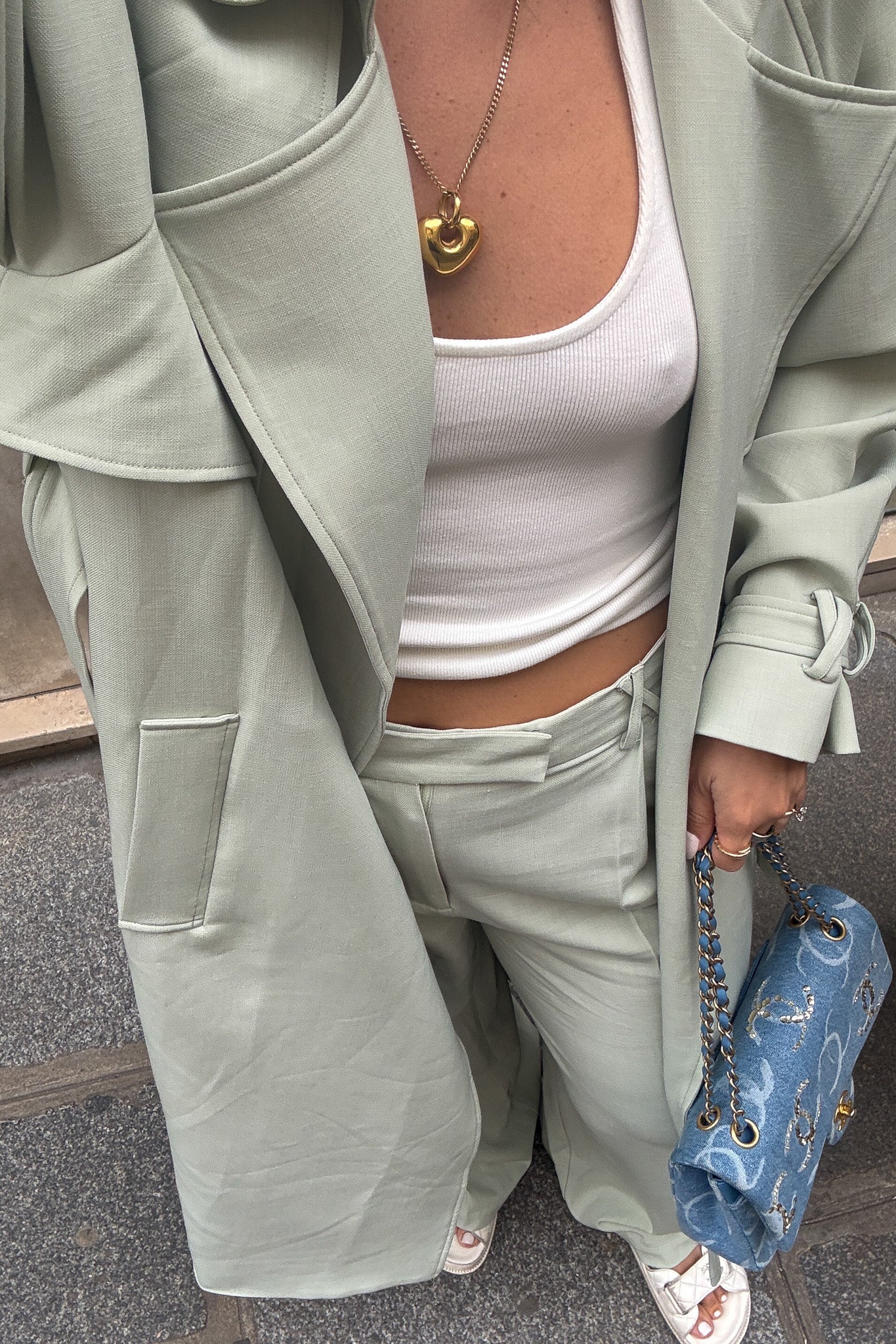 Woman in a light green coat and pants standing on a sidewalk with a building and hedge in the background